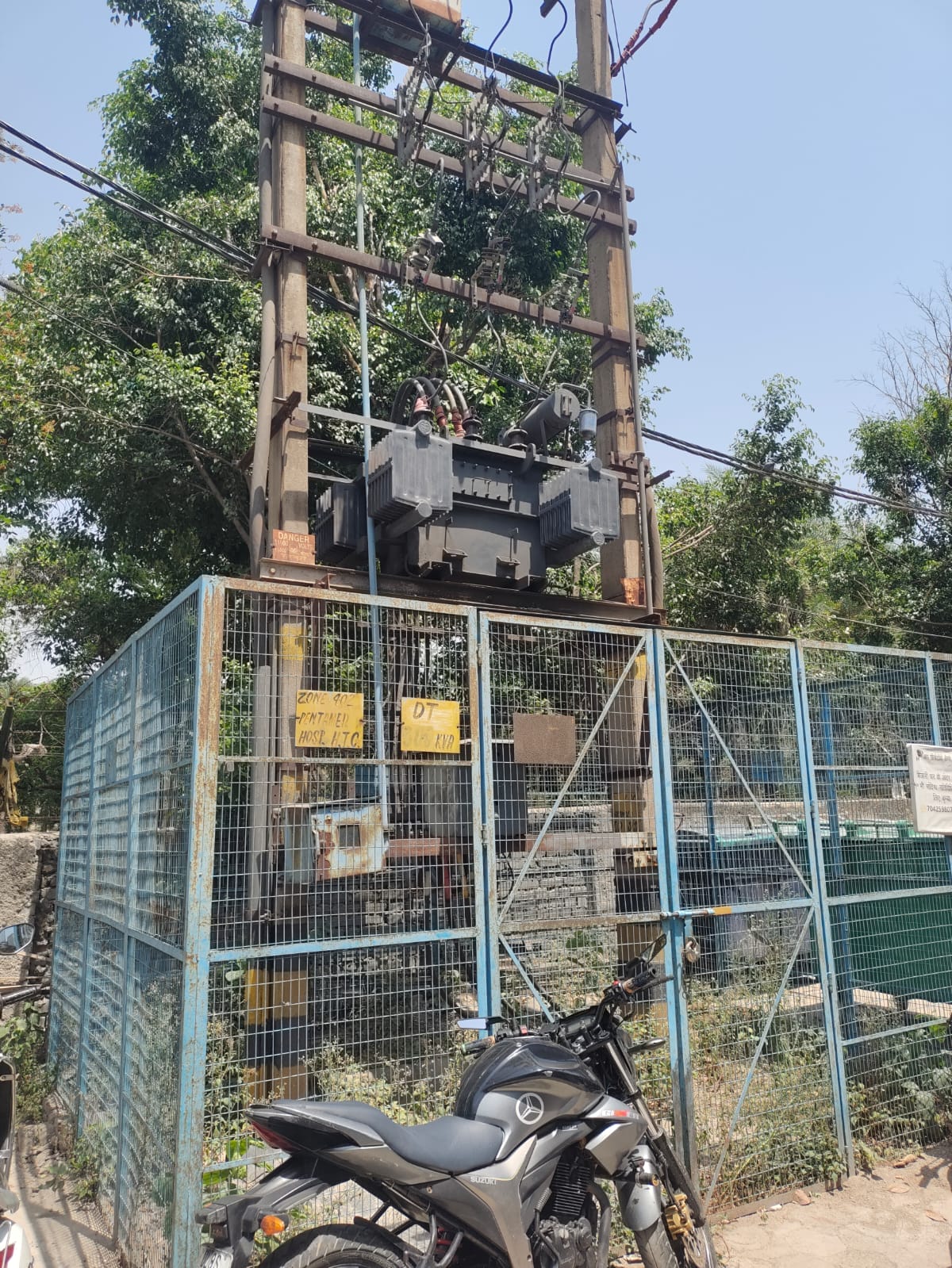 A large electrical transformer is mounted on a metal structure and enclosed by a blue wire fence. Warning signs promoting electrical safety compliance are posted, with trees in the background and a parked motorcycle in the foreground.
