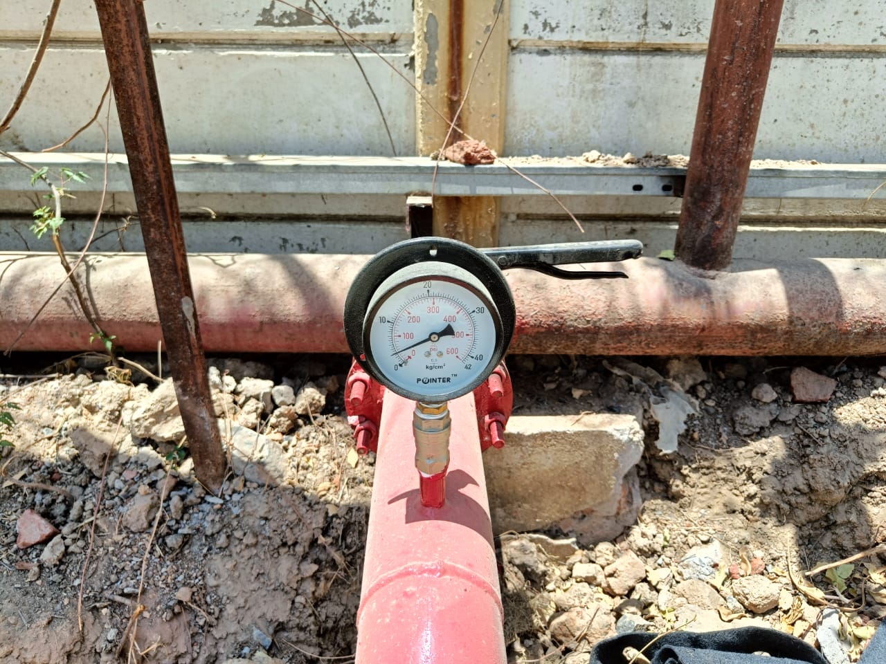 A close-up of a pressure gauge attached to a red pipeline outdoors, surrounded by dry soil, rocks, and metal pipes—a typical scene during a fire safety inspection or fire risk assessment, with the gauge needle pointing to a reading against a concrete wall.