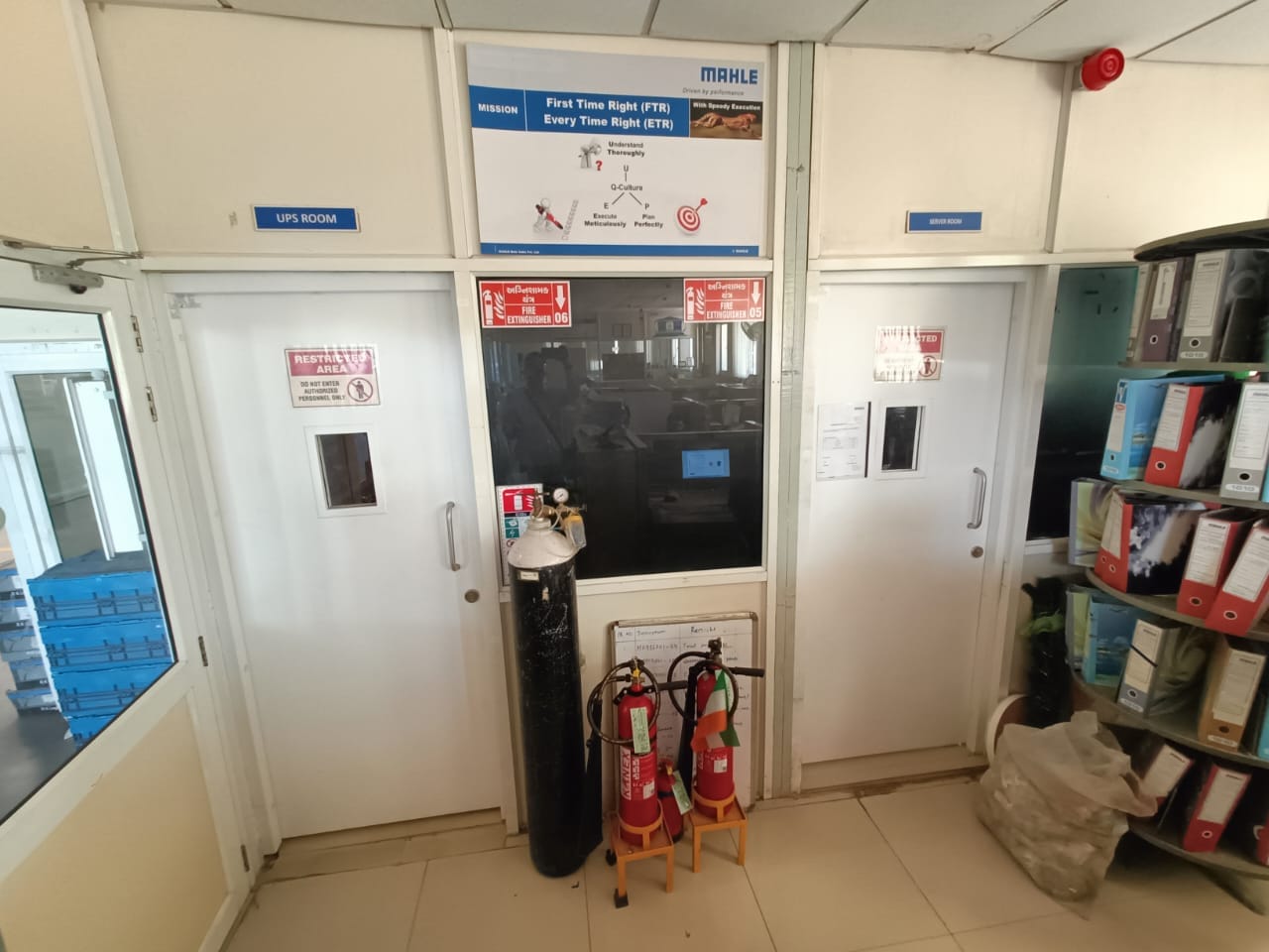 A room with two white doors labeled UPS Room and Server Room, fire extinguishers, an oxygen cylinder, safety signs, and shelves with files and bags—ideal for a fire safety audit. A sign above the doors reads First Time Right, Every Time Right.