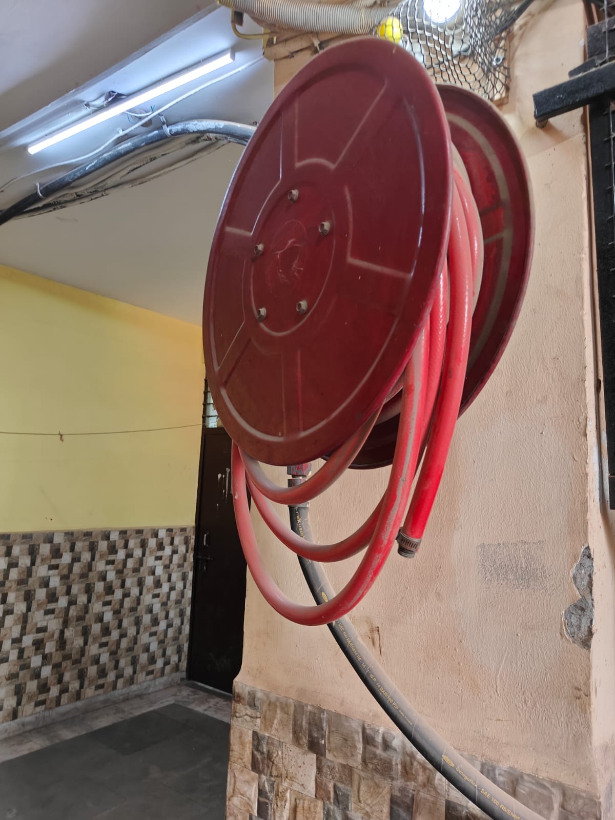 A red fire hose reel is mounted on a beige wall in a hallway with tiled and painted surfaces, illuminated by overhead fluorescent lights—a crucial feature highlighted during any thorough fire safety inspection.