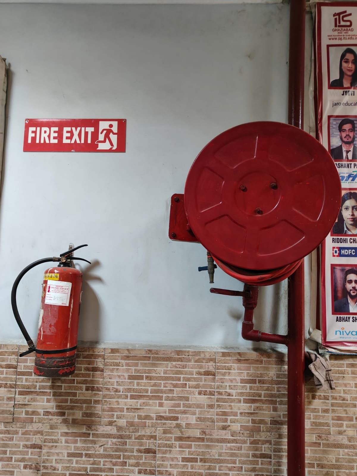 A red fire extinguisher and fire hose reel are mounted on a tiled wall under a red Fire Exit sign, beside photo posters—an ideal setup to feature in a fire audit for safety compliance.