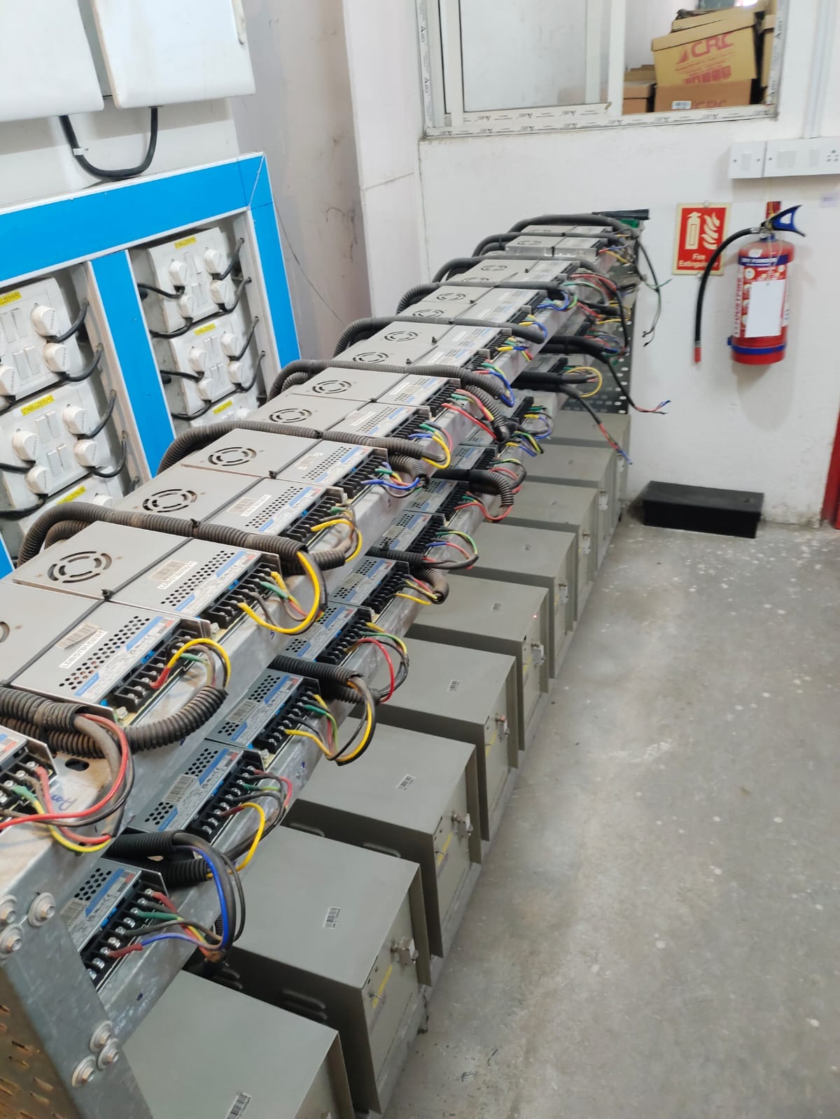 A row of electrical equipment and power supplies with multiple cables connected, installed on a metal rack in an industrial room during machinery vibration testing. A fire extinguisher is mounted on the wall nearby for industrial maintenance safety.