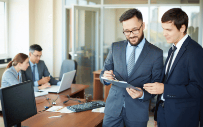 Two men in business suits discuss documents on a clipboard in an office, while a man and woman work at computers in the background. The setting is a professional, modern workspace.