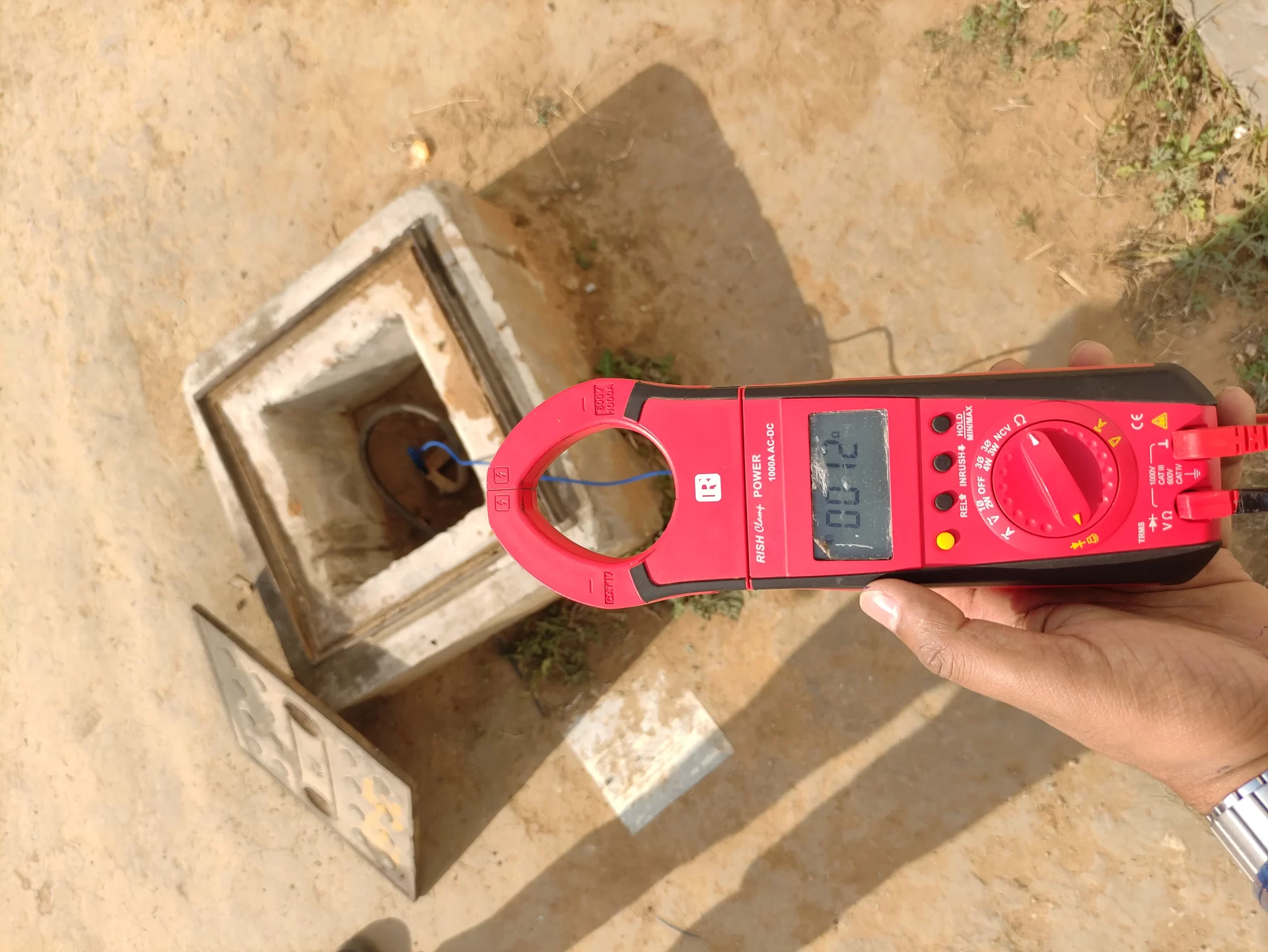 A person holds a red digital clamp meter displaying 0.12 near an open ground electrical box with exposed wires on a dusty outdoor surface, part of an Elion sustainability audit.