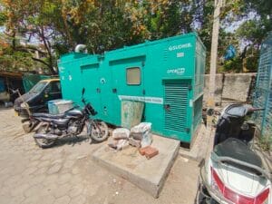 A turquoise Sudhir generator sits outdoors on a concrete base beside parked motorcycles at a dairy unit in Anand, Gujarat, surrounded by trees and construction materials in a sunny urban setting—ideal for an Elion energy audit.