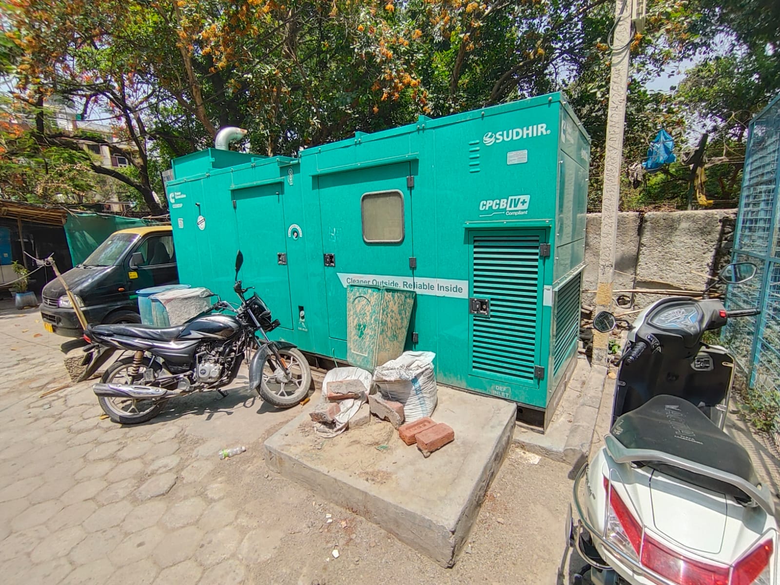 A turquoise Sudhir generator sits outdoors on a concrete base beside parked motorcycles at a dairy unit in Anand, Gujarat, surrounded by trees and construction materials in a sunny urban setting—ideal for an Elion energy audit.