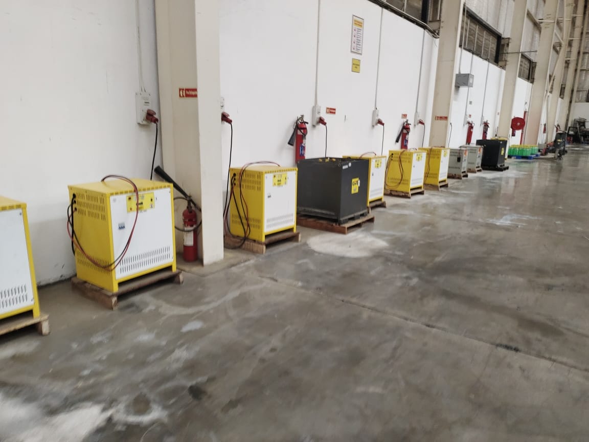 Several yellow and gray industrial battery chargers are arranged on wooden pallets along a warehouse wall in an IT park, Bengaluru, Karnataka, with fire extinguishers mounted between each unit to support safety awareness and fire prevention.