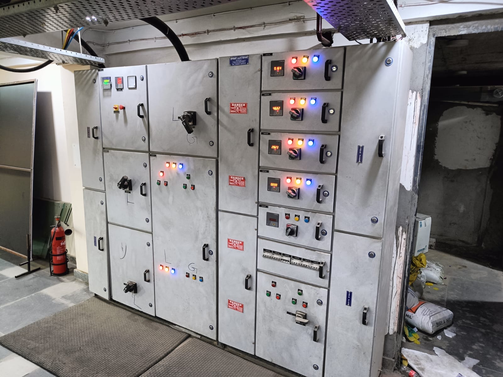 Large industrial electrical control panel with multiple switches, indicator lights, and meters, situated in a utility room of a textile mill in Coimbatore. A fire extinguisher and cleaning supplies nearby highlight attention to industrial electrical safety.