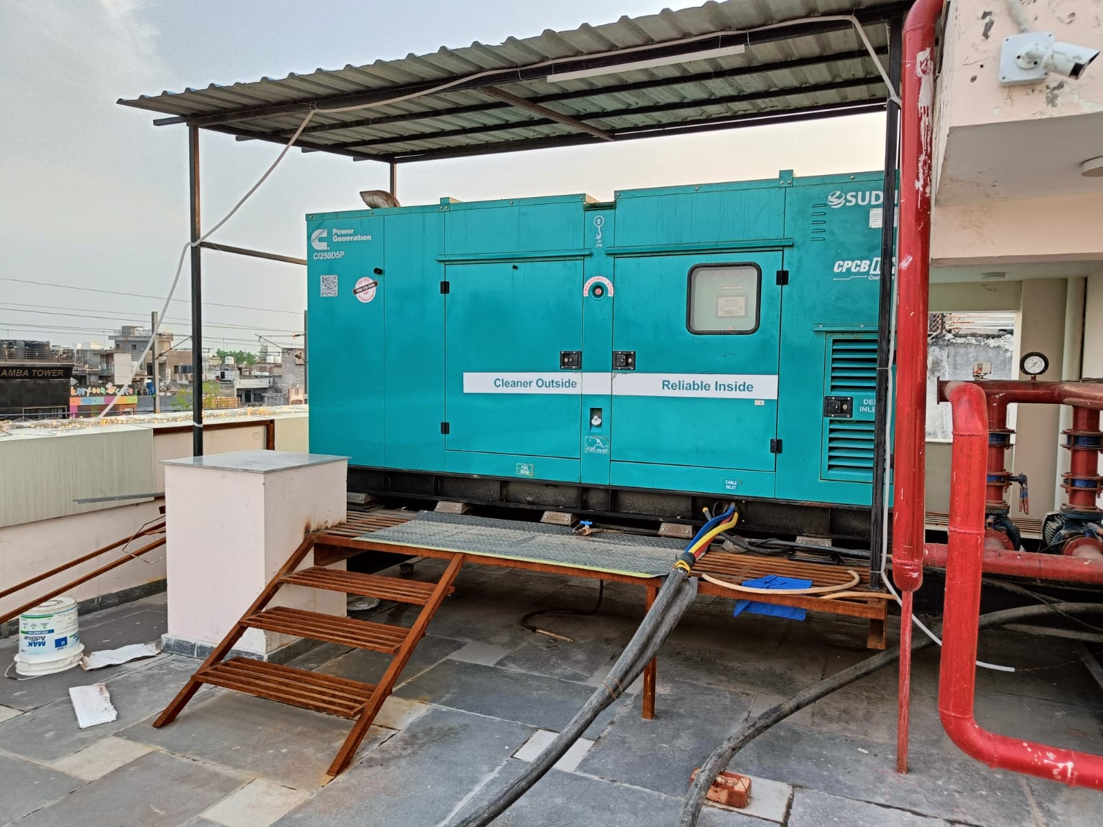 A large teal industrial generator with Cleaner Outside Reliable Inside written on it is installed on a rooftop in Tirupur, supporting a textile manufacturer’s industrial energy saving efforts, with cables, pipes, and wooden ramps connected under a metal canopy.
