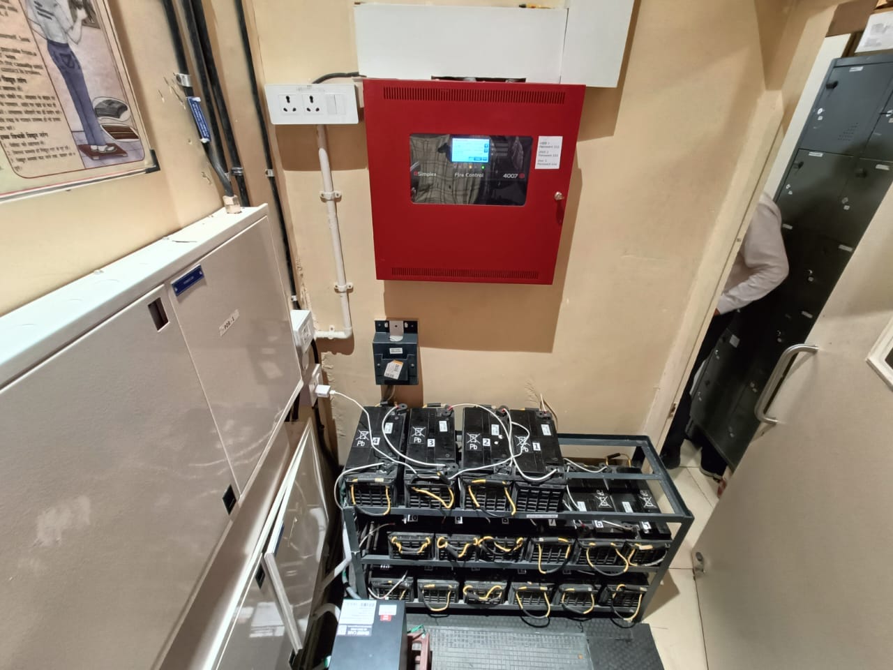 A small room in a Raipur steel foundry with electrical equipment, including a red control panel, battery rack with cables, and a white panel. An Elion industrial inspection or electrical safety audit may be underway as a person stands at the open door.