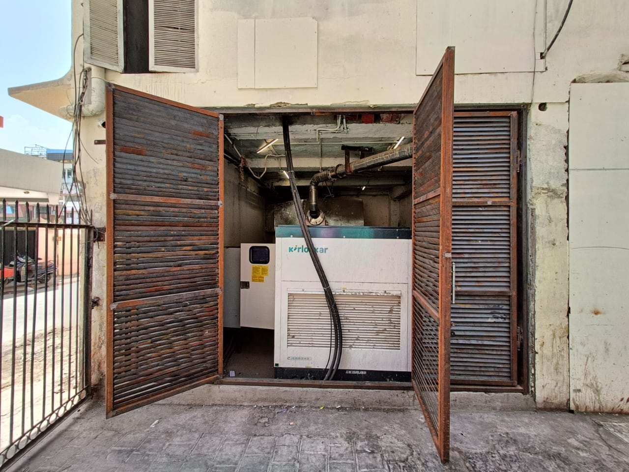 A large white industrial generator labeled “Kirloskar” is placed inside a small concrete room with both rusty metal doors open. Black pipes and cables are connected to the generator.