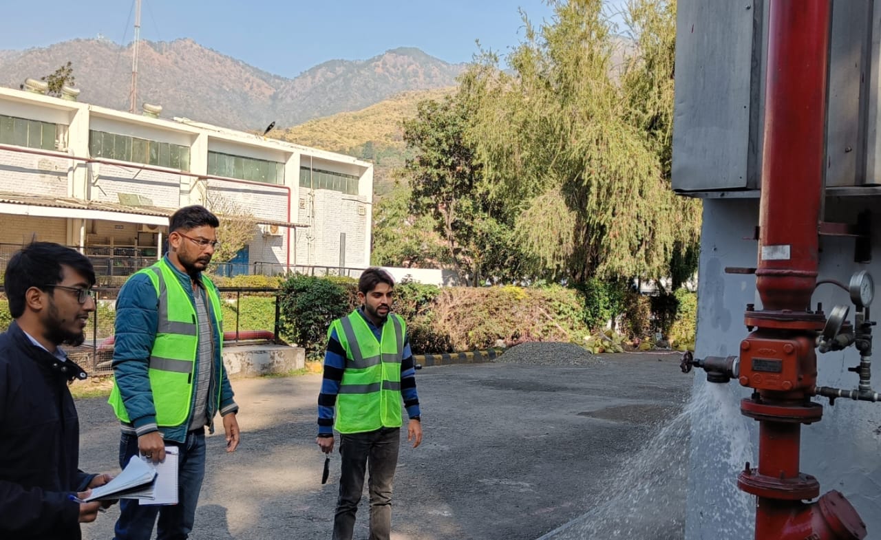 Three men in green safety vests stand outdoors near a red industrial pipe, with one holding a notepad and referencing the Lift Safety Audit Checklist. A building, greenery, and mountains appear in the background under a clear sky.