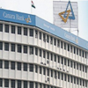 A multi-story white building with Canara Bank signage and logo on the facade and rooftop, along with the Indian flag flying above.