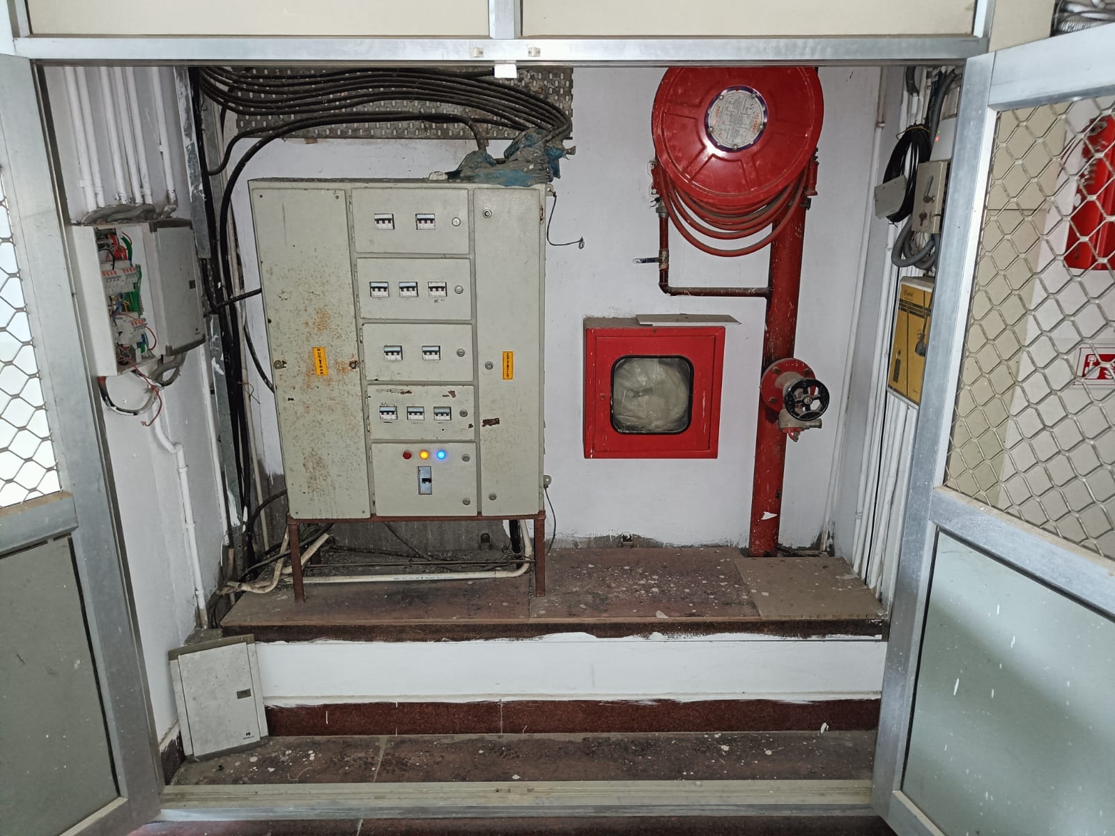 An electrical control panel with multiple switches sits next to a red fire hose reel and fire extinguisher behind a metal fence in a building utility area. The wall and floor show signs of dust and wear.