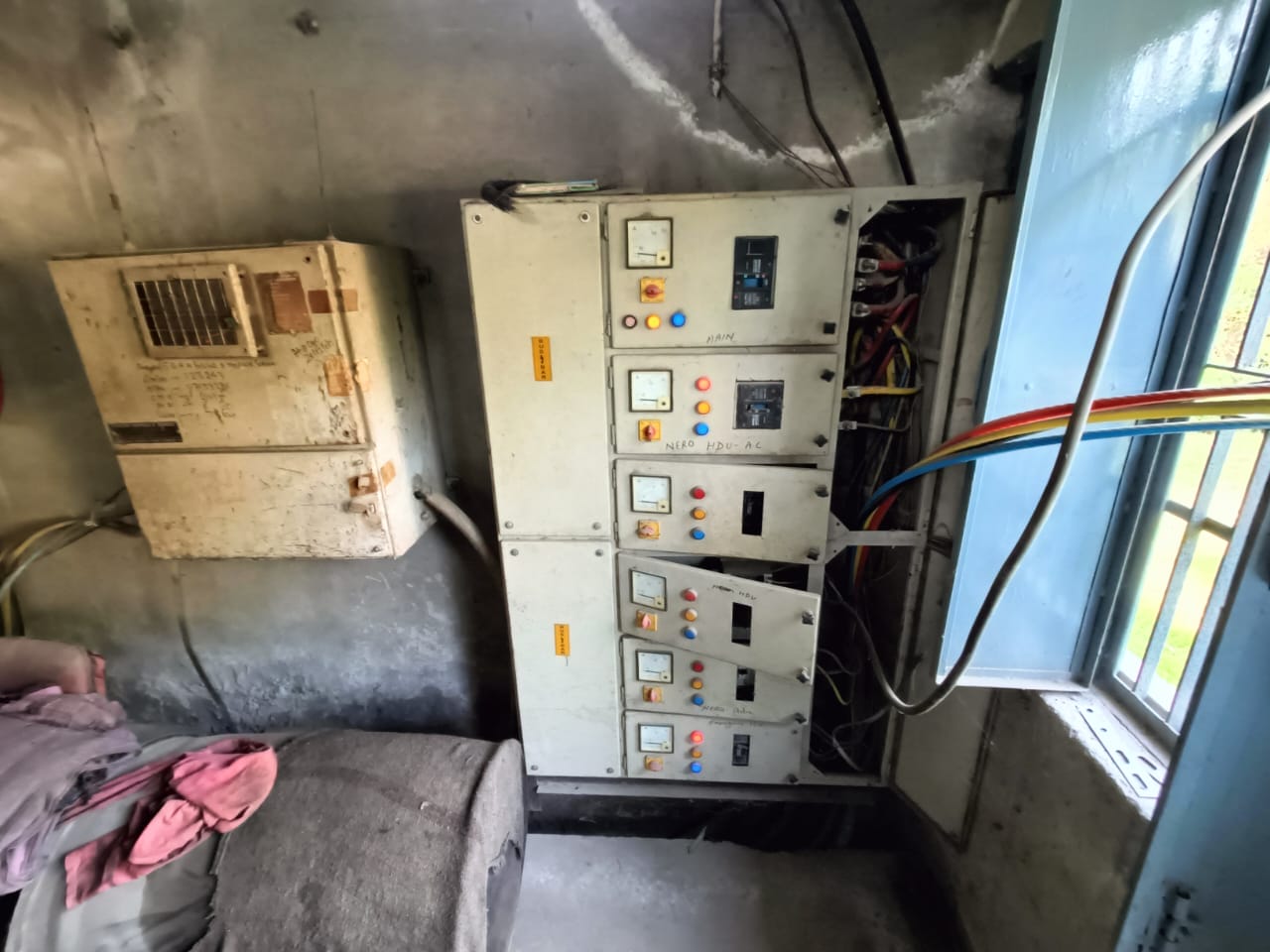 A room with electrical panels mounted on a wall, showing switches, meters, and exposed wires. A window is open to the right, and a dirty cloth and a large object are visible on the floor to the left.