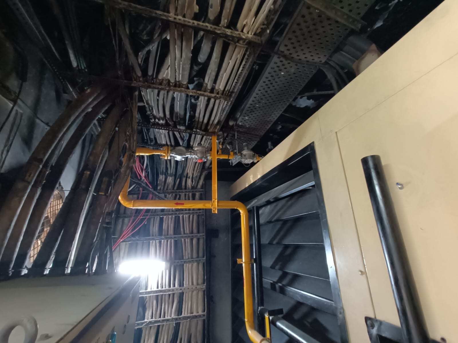 View of an industrial ceiling showing exposed electrical cables, a yellow metal gas pipeline with valves, metal beams, and a partially open black ventilation grille on the right—ideal for conducting an energy audit by Elion services for industries in India.