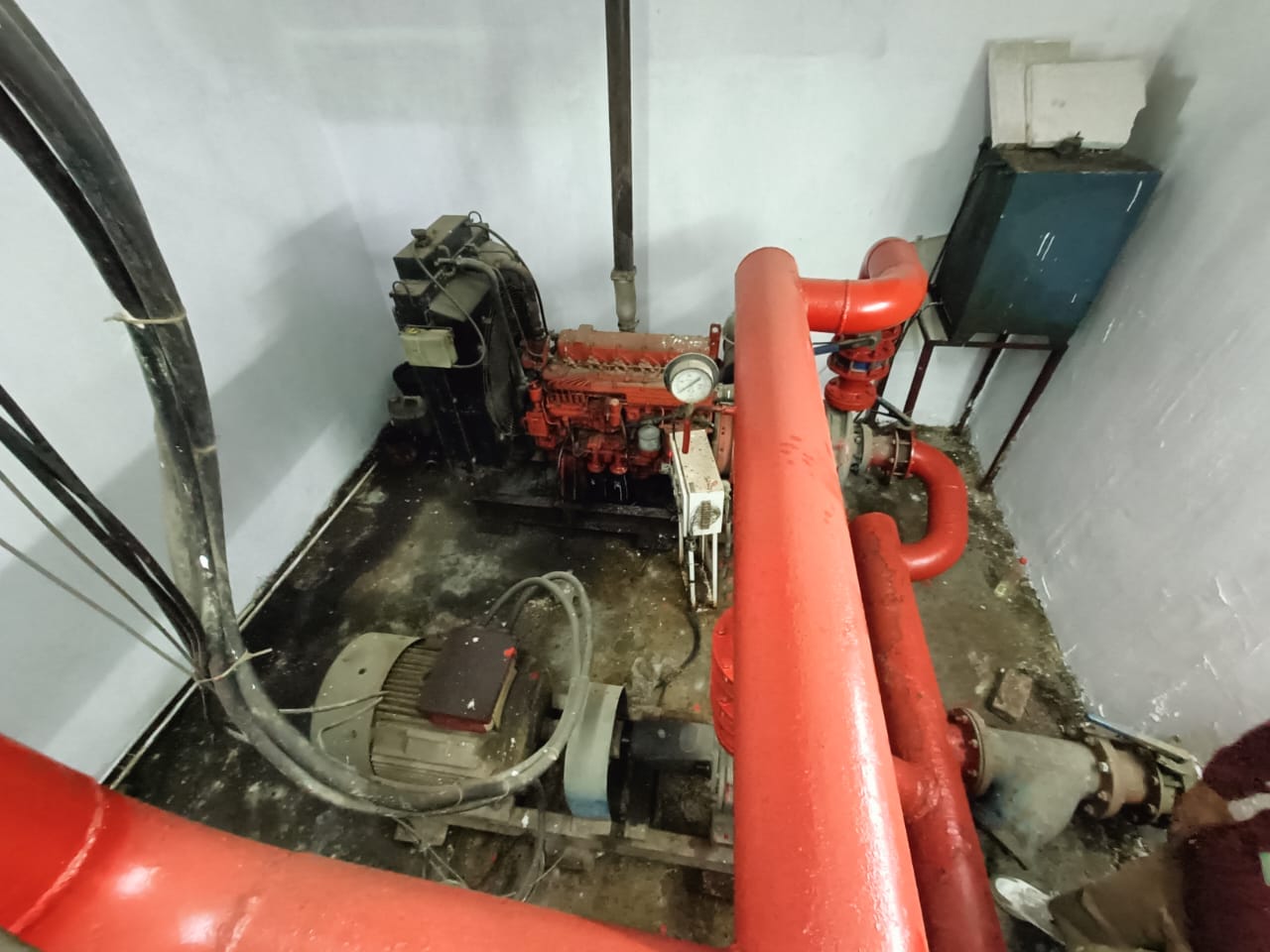 A room with industrial equipment, including a red pipe system, a pump, and a control panel mounted on the wall. The floor appears worn and the setting looks like a utility or mechanical room.