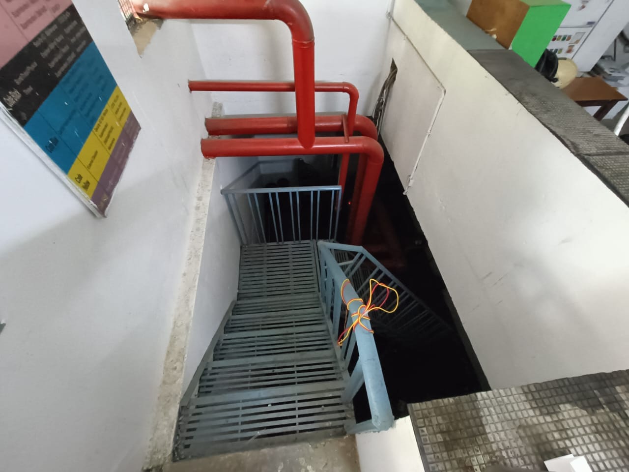 A metal staircase leads downwards in a narrow indoor space with white walls and red pipes overhead. An orange cable is tied to the blue railing, and nearby are colorful signs and office furniture.