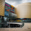A modern shopping mall entrance with gold, perforated walls, large glass windows, and visible store signs including Reserved, CCC, and others. People are walking near the entrance under a cloudy sky.