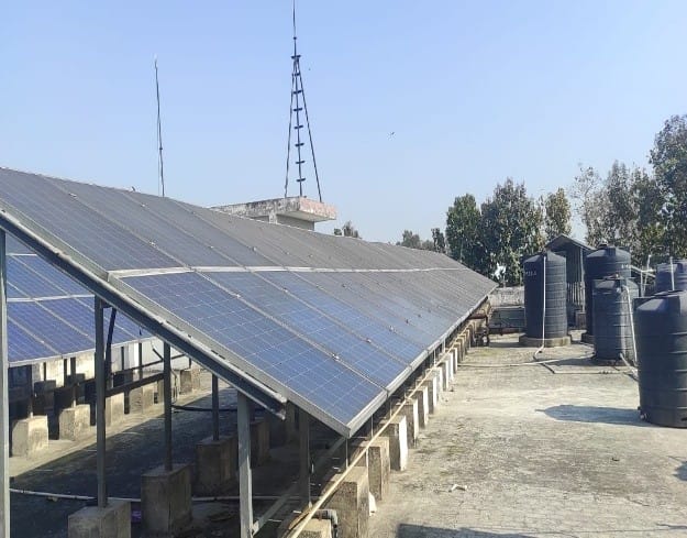 Solar panels installed on a flat rooftop with several black water storage tanks and trees in the background under a clear blue sky.