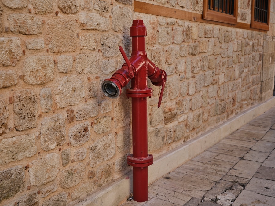A red fire hydrant is attached to a stone wall on a paved sidewalk, with two side valves and an open connector facing forward.