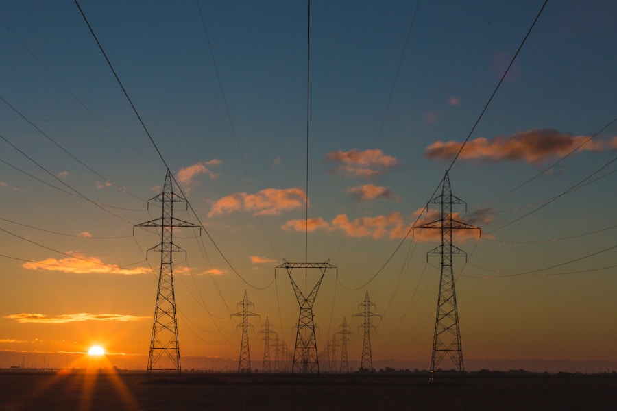 High-voltage power lines and pylons stretch across a flat landscape at sunset, with the sun low on the horizon and a colorful sky with scattered clouds.