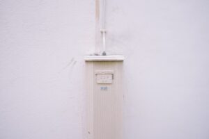 A beige utility box mounted on a plain, light-colored wall with a thin pipe extending upward from the box. The wall and box are clean, and the scene appears minimalistic and uncluttered.