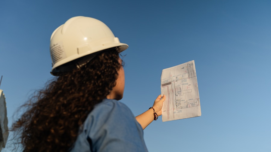 A person wearing a white safety helmet holds up and examines a technical blueprint or construction plan, standing outdoors under a clear blue sky.