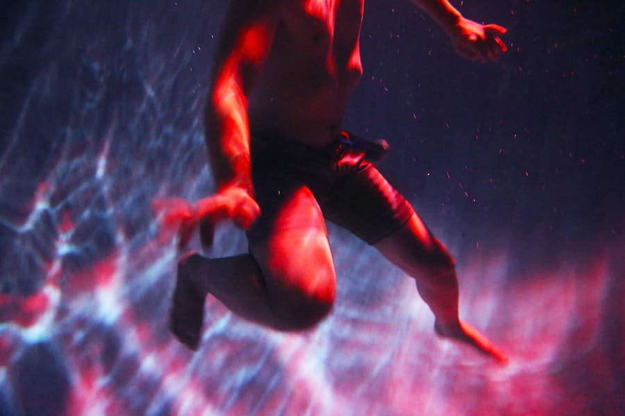 A person in swim shorts is underwater, illuminated by dramatic red and blue lighting, with arms and legs bent as if floating or swimming. Rippling light patterns are visible on the water around them.
