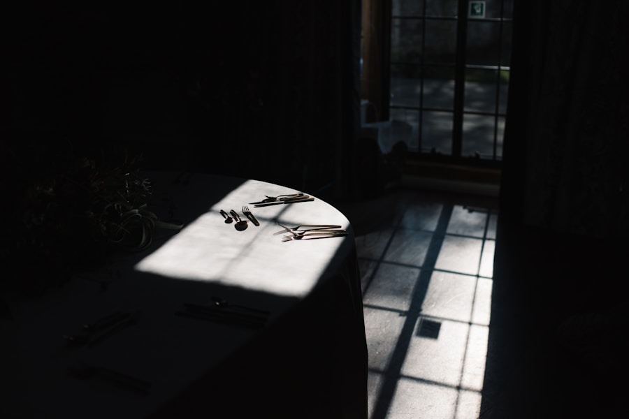 A round table with cutlery is partially illuminated by sunlight streaming through a window, casting shadows across the floor in an otherwise dimly lit room.