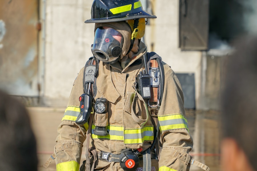 A firefighter in full protective gear and breathing mask stands outdoors, with a radio and safety equipment attached to their uniform. The background shows a blurry building structure.