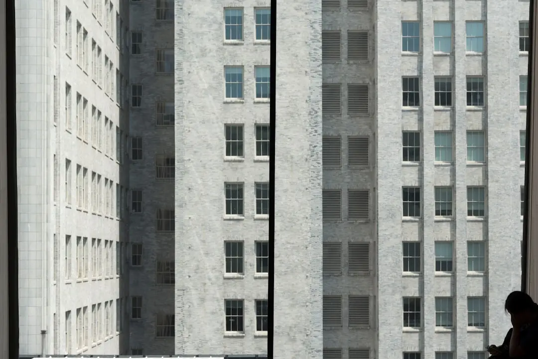 A person stands near a window in silhouette, looking out at a tall, gray brick building with many windows. The scene is divided by a vertical window frame.