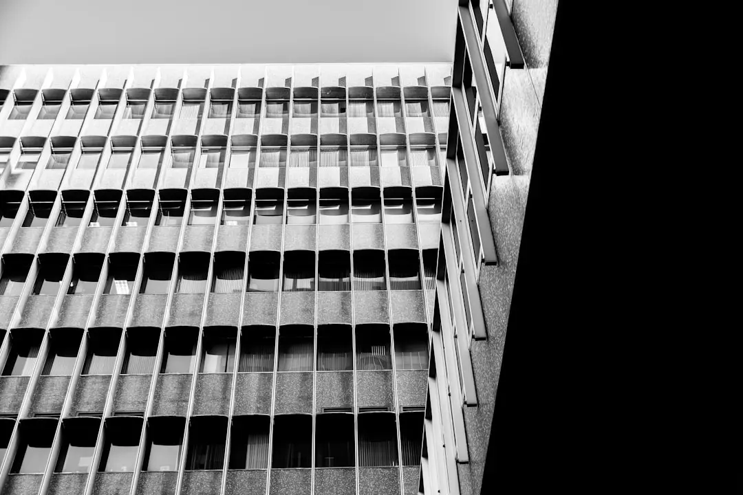 Black and white photo of a modern office building with repetitive window patterns and concrete panels, seen from a low angle. Another building edge frames the right side of the image.