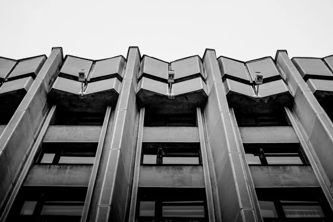 Black and white photo of a tall, concrete building with a geometric, angular facade. The perspective is from below, highlighting the repetitive patterns and architectural details against a clear sky.
