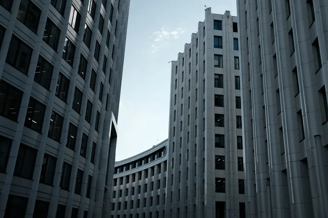 Tall, modern office buildings with many windows create a narrow corridor, leading to a curved building in the background, against a partly cloudy sky.