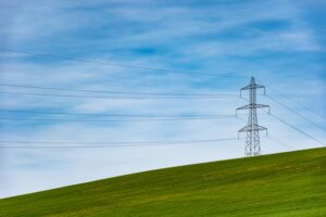 A metal electricity pylon stands on a grassy hill under a blue sky with wispy clouds, with power lines stretching across the scene.