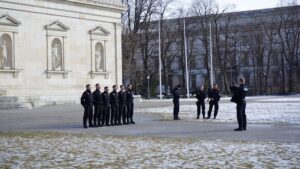 A group of uniformed officers stand in a line facing an officer, with a few more people gathered nearby in an outdoor area with patches of snow and historic buildings in the background.