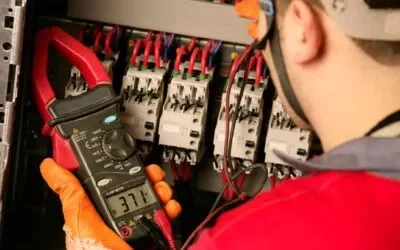An electrician wearing orange gloves uses a digital clamp meter to measure voltage or current in an electrical panel with multiple wires, performing tasks essential for a thorough Energy Audit.