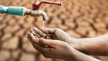 A pair of dirty hands are held under a dripping faucet, with only a few drops of water falling, set against a background of dry, cracked earth, symbolizing water scarcity and drought.